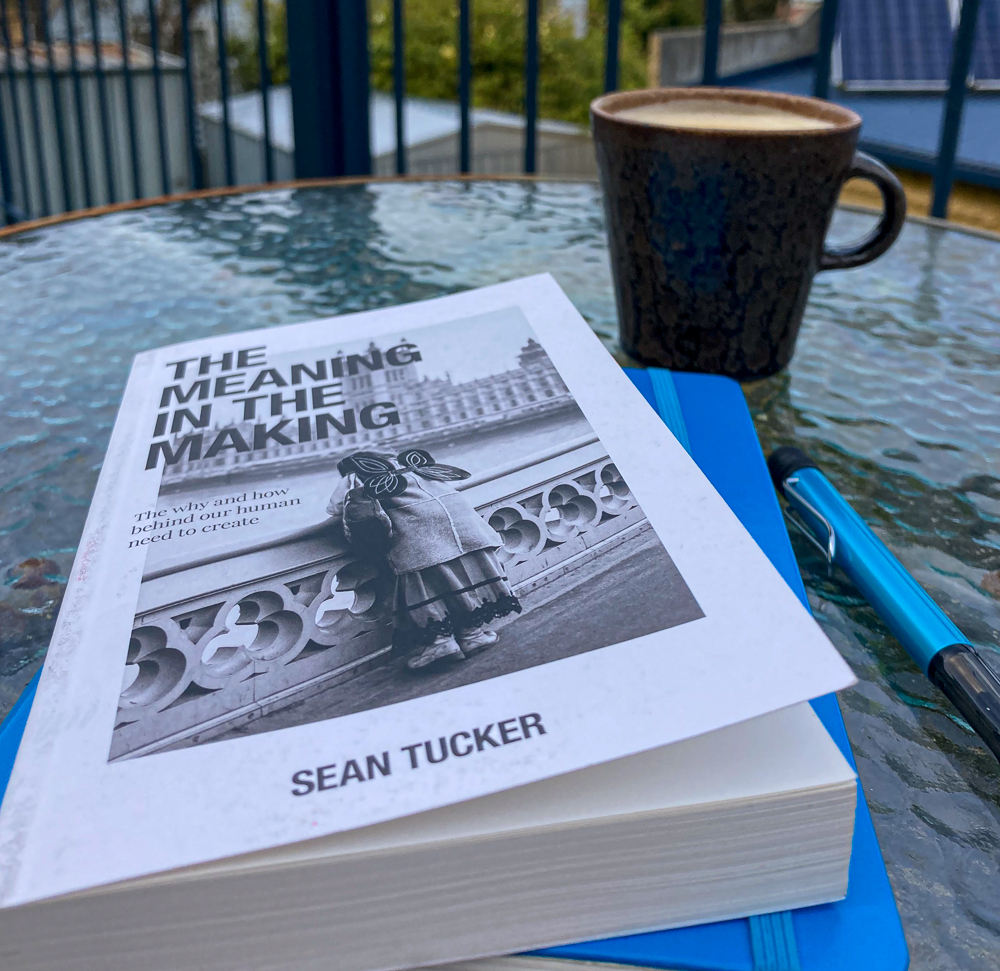 A copy of Sean Tucker's book The Meaning in the Making on a table with a blue notebook, a blue pen and a cup of coffee