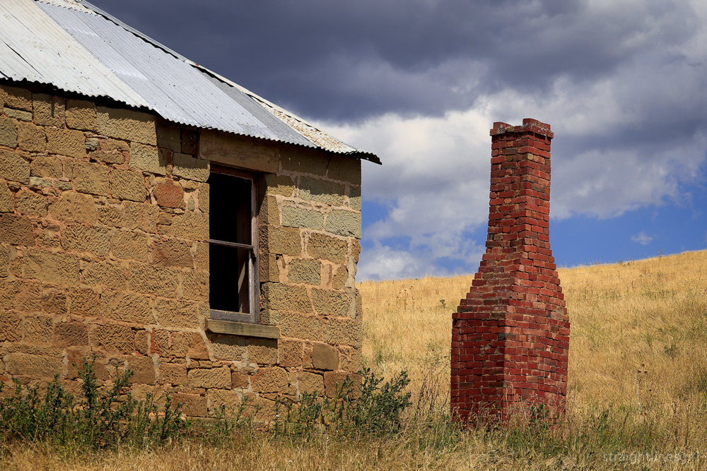The side of an old sandstone cottage, a red brick chimney in a field of dry grass and dark clouds overhead